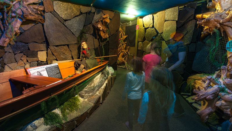 Een museumexpositie met een boot, een heksenpop, stenen muren en wazige bezoekers.